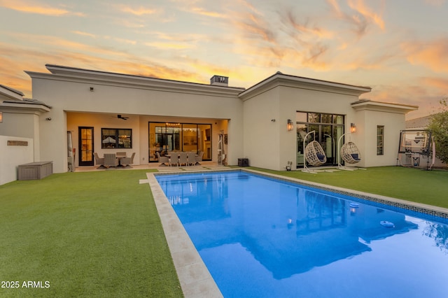 back of property at dusk with stucco siding, an outdoor pool, a lawn, and a patio