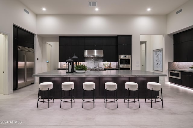 kitchen with tasteful backsplash, a kitchen bar, built in appliances, and a large island with sink