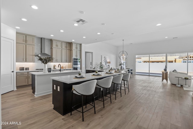 kitchen with an island with sink, light hardwood / wood-style floors, wall chimney range hood, a kitchen bar, and decorative light fixtures