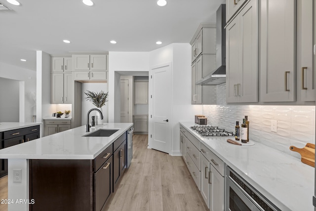 kitchen with an island with sink, sink, wall chimney range hood, appliances with stainless steel finishes, and light hardwood / wood-style floors