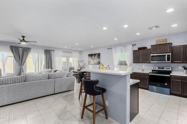 kitchen with a kitchen bar, dark brown cabinetry, stainless steel appliances, a center island with sink, and plenty of natural light