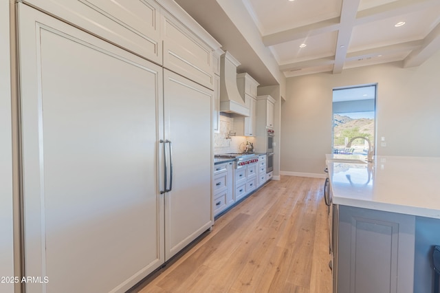 kitchen with coffered ceiling, custom exhaust hood, white cabinetry, light hardwood / wood-style flooring, and backsplash