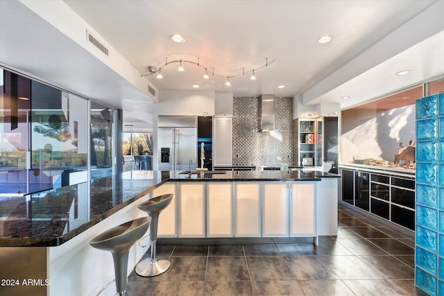 kitchen with backsplash, wall chimney exhaust hood, sink, dark stone countertops, and built in fridge