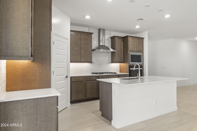 kitchen with dark brown cabinetry, sink, stainless steel appliances, wall chimney range hood, and a center island with sink