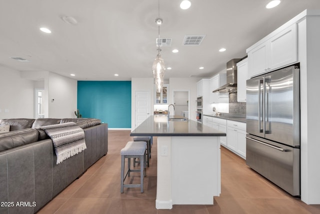 kitchen with white cabinetry, wall chimney range hood, hanging light fixtures, stainless steel appliances, and a center island with sink