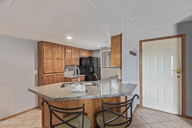 kitchen with a sink, black fridge, light tile patterned flooring, brown cabinetry, and a peninsula