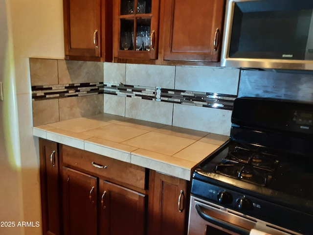 kitchen featuring gas range oven, backsplash, tile counters, stainless steel microwave, and glass insert cabinets