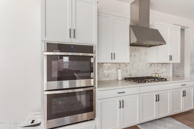 kitchen with light countertops, appliances with stainless steel finishes, white cabinetry, wall chimney exhaust hood, and backsplash