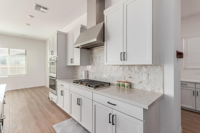 kitchen featuring visible vents, stainless steel appliances, light wood-style floors, wall chimney exhaust hood, and decorative backsplash