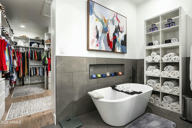 bathroom with a freestanding tub, a wainscoted wall, wood finished floors, tile walls, and a walk in closet