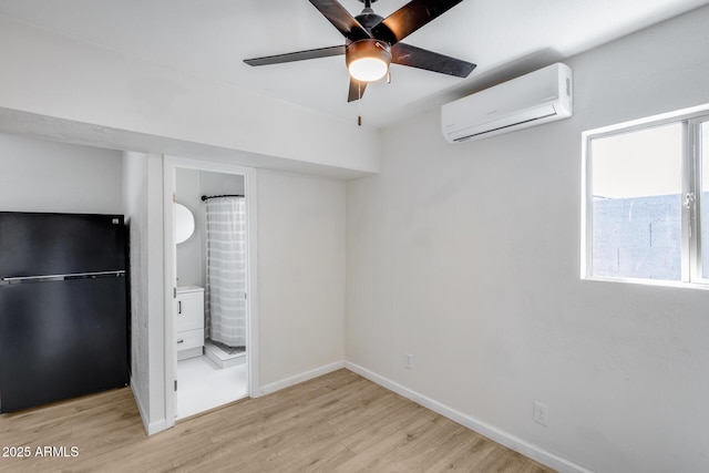unfurnished bedroom featuring baseboards, an AC wall unit, light wood-type flooring, freestanding refrigerator, and ensuite bath