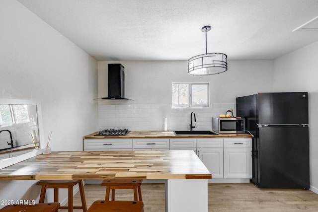 kitchen featuring wall chimney range hood, a sink, stainless steel microwave, freestanding refrigerator, and butcher block counters