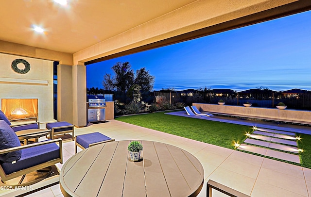 patio terrace at dusk with area for grilling, a yard, an outdoor fireplace, and an outdoor kitchen