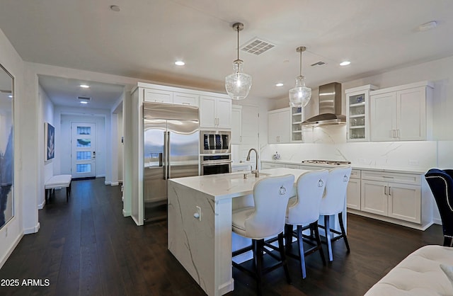 kitchen with hanging light fixtures, built in appliances, white cabinets, a center island with sink, and wall chimney exhaust hood