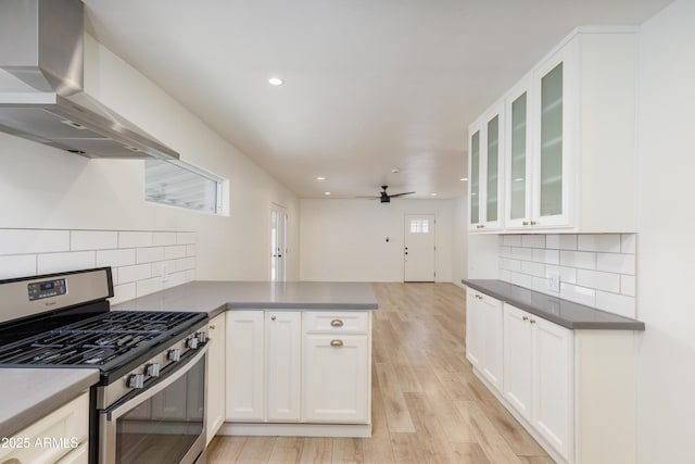 kitchen featuring light hardwood / wood-style flooring, stainless steel range with gas cooktop, kitchen peninsula, wall chimney range hood, and white cabinets