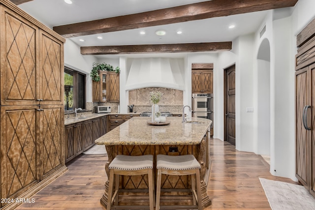 kitchen with hardwood / wood-style floors, custom exhaust hood, an island with sink, and tasteful backsplash