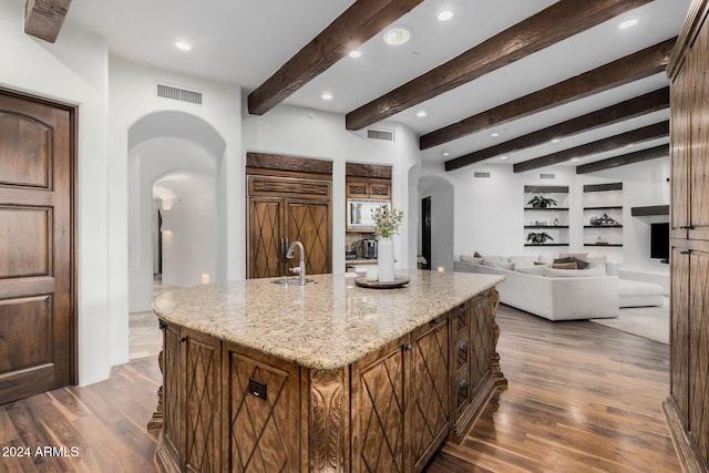 kitchen featuring light stone countertops, beamed ceiling, dark hardwood / wood-style floors, paneled built in fridge, and a center island with sink