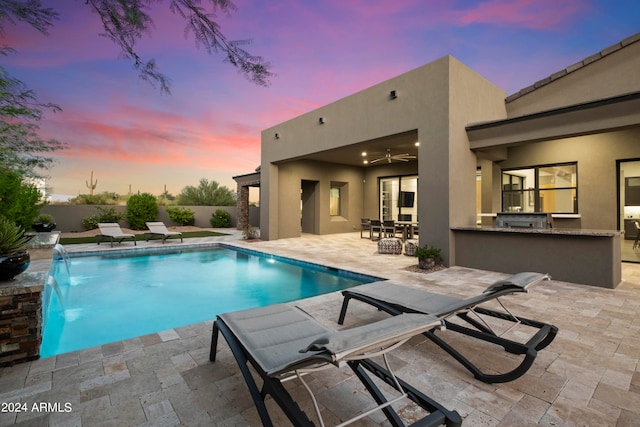 pool at dusk with pool water feature, a patio area, a bar, ceiling fan, and exterior kitchen