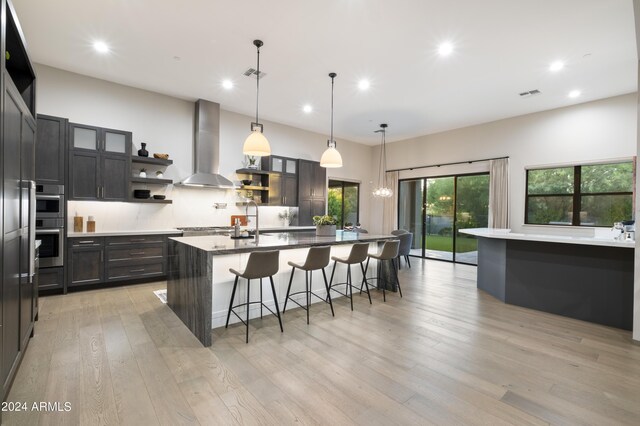 kitchen featuring decorative light fixtures, a breakfast bar area, wall chimney exhaust hood, a center island with sink, and light hardwood / wood-style flooring