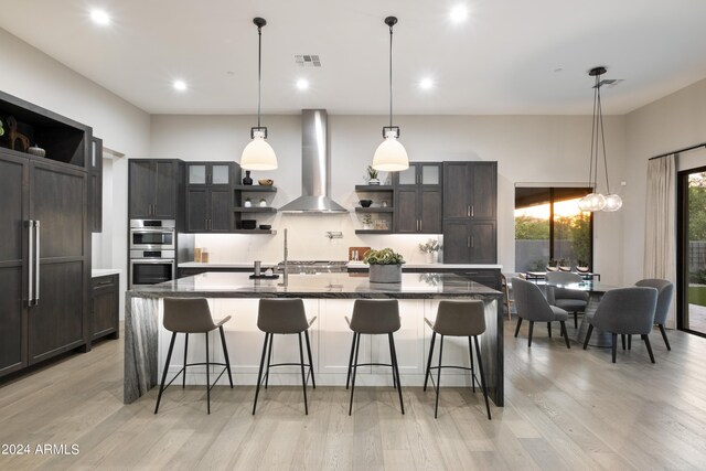 kitchen with a breakfast bar, a kitchen island with sink, stainless steel appliances, dark brown cabinets, and wall chimney exhaust hood