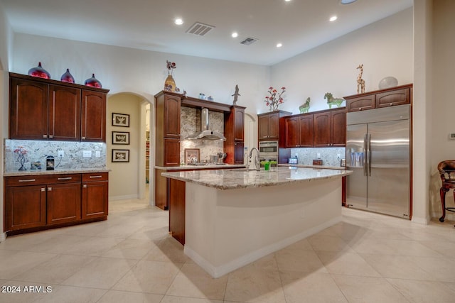 kitchen with wall chimney range hood, tasteful backsplash, built in fridge, light stone counters, and a center island with sink