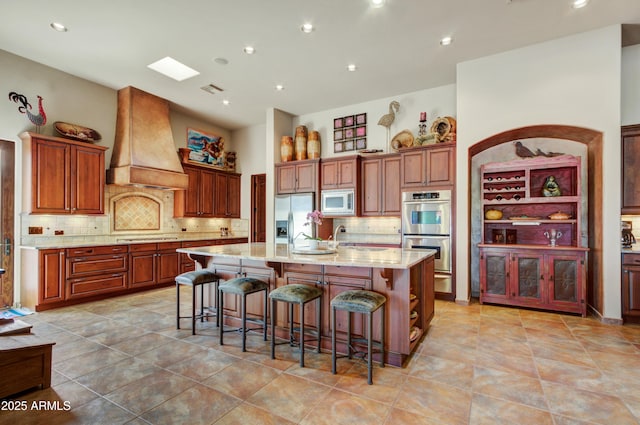 kitchen with appliances with stainless steel finishes, a kitchen bar, custom exhaust hood, light stone countertops, and a center island with sink