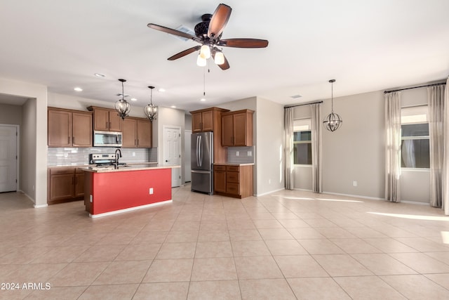 kitchen with tasteful backsplash, ceiling fan with notable chandelier, stainless steel appliances, pendant lighting, and an island with sink