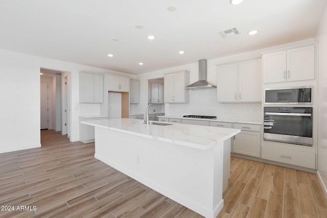 kitchen featuring wall chimney exhaust hood, stainless steel appliances, a kitchen island with sink, light hardwood / wood-style floors, and white cabinetry