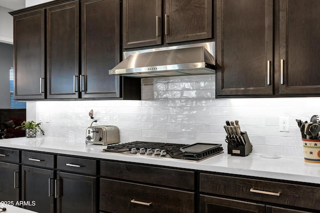 kitchen featuring dark brown cabinetry, light countertops, under cabinet range hood, stainless steel gas stovetop, and backsplash