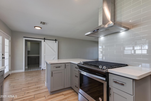 kitchen featuring light countertops, stainless steel gas stove, a barn door, wall chimney exhaust hood, and gray cabinetry