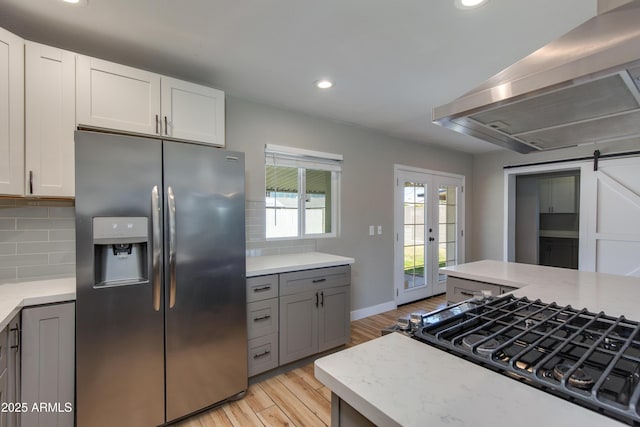kitchen featuring light wood-type flooring, stainless steel fridge with ice dispenser, decorative backsplash, a barn door, and wall chimney range hood