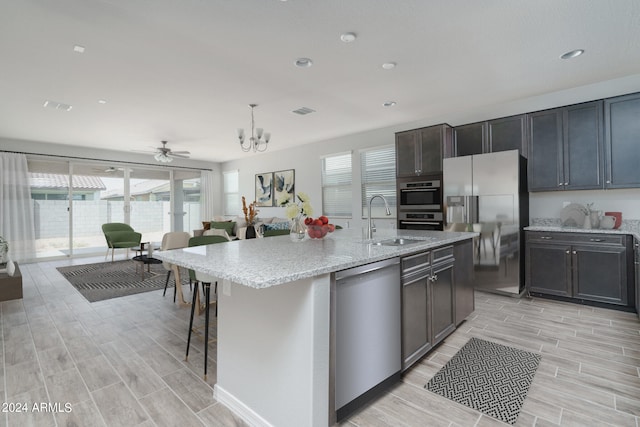 kitchen with light stone counters, ceiling fan with notable chandelier, stainless steel appliances, sink, and an island with sink