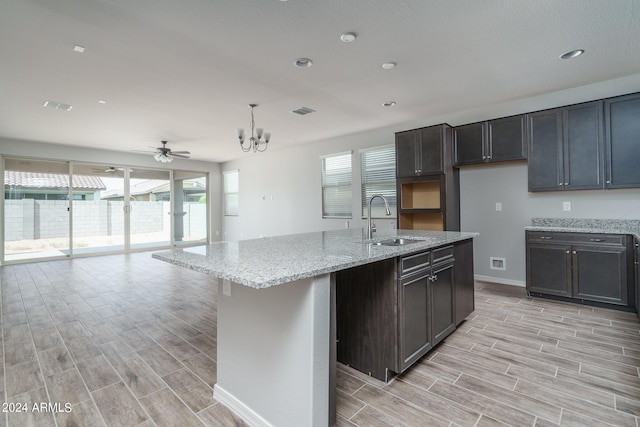 kitchen featuring light stone countertops, ceiling fan with notable chandelier, a kitchen island with sink, sink, and hanging light fixtures