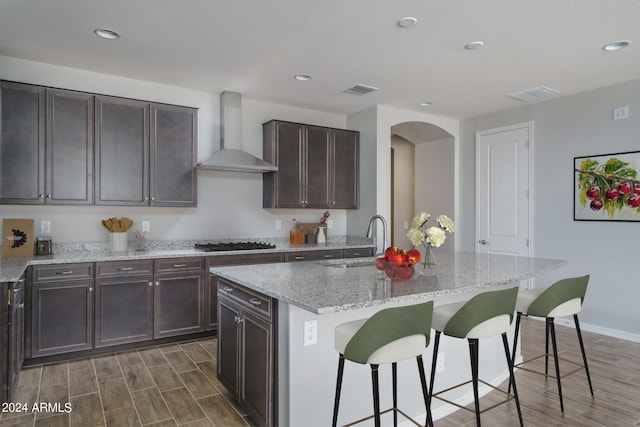 kitchen with a kitchen island with sink, a kitchen breakfast bar, sink, wall chimney exhaust hood, and stainless steel gas cooktop