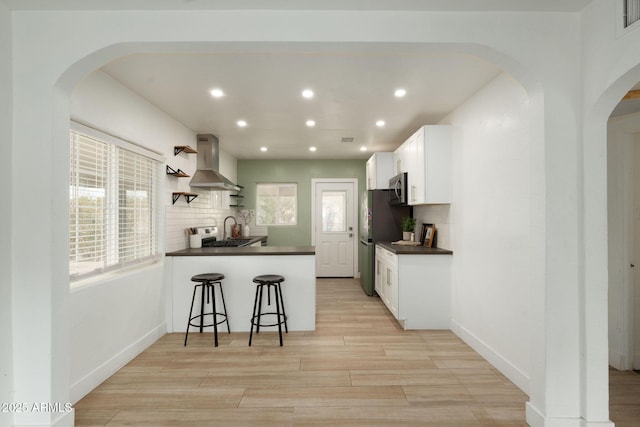 kitchen featuring arched walkways, dark countertops, appliances with stainless steel finishes, and wall chimney exhaust hood