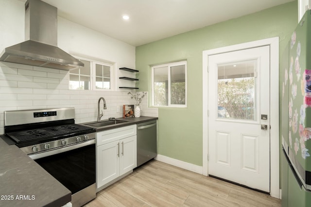 kitchen featuring backsplash, wall chimney range hood, appliances with stainless steel finishes, white cabinets, and a sink