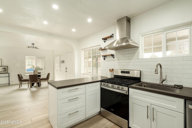 kitchen with dark countertops, wall chimney range hood, a peninsula, stainless steel gas stove, and a sink