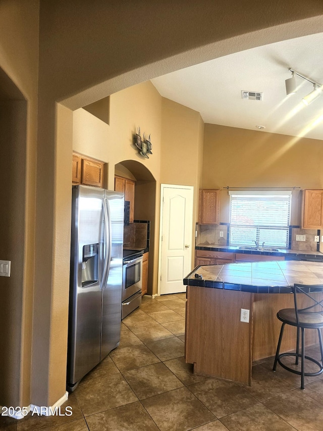 kitchen with tile countertops, vaulted ceiling, a kitchen breakfast bar, stainless steel appliances, and backsplash