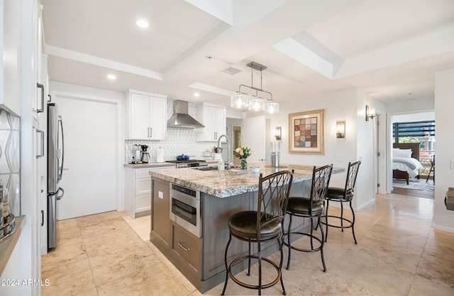 kitchen featuring light stone countertops, a kitchen island with sink, wall chimney range hood, decorative light fixtures, and white cabinetry