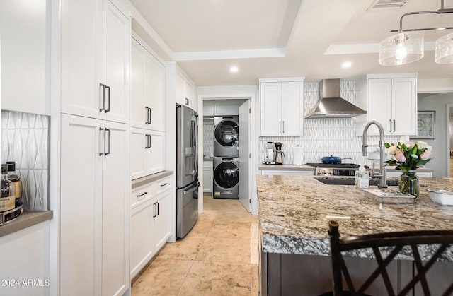 kitchen featuring white cabinetry, wall chimney exhaust hood, hanging light fixtures, light stone counters, and stacked washer and clothes dryer