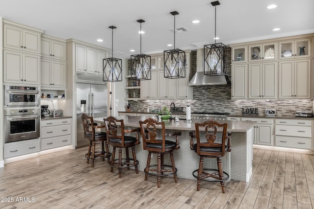 kitchen featuring wall chimney exhaust hood, a center island with sink, pendant lighting, stainless steel appliances, and cream cabinets