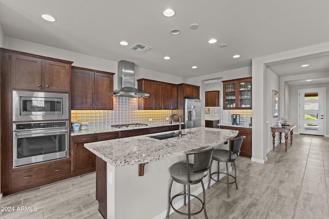 kitchen featuring appliances with stainless steel finishes, a kitchen island with sink, sink, wall chimney exhaust hood, and light stone counters