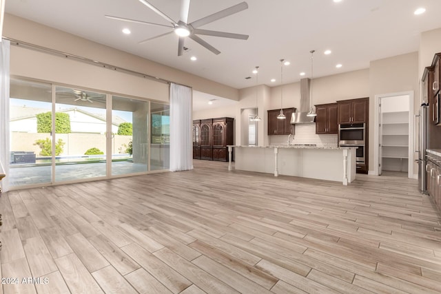 kitchen featuring wall chimney range hood, decorative backsplash, light wood-type flooring, light stone counters, and stainless steel double oven