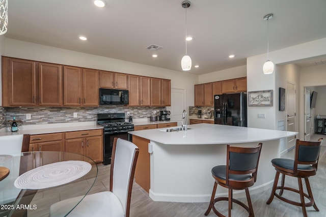 kitchen featuring sink, backsplash, hanging light fixtures, black appliances, and an island with sink