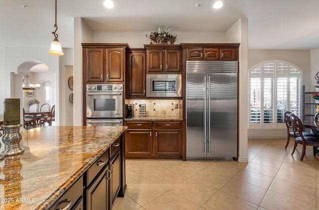 kitchen with tasteful backsplash, light tile patterned flooring, hanging light fixtures, built in appliances, and stone counters