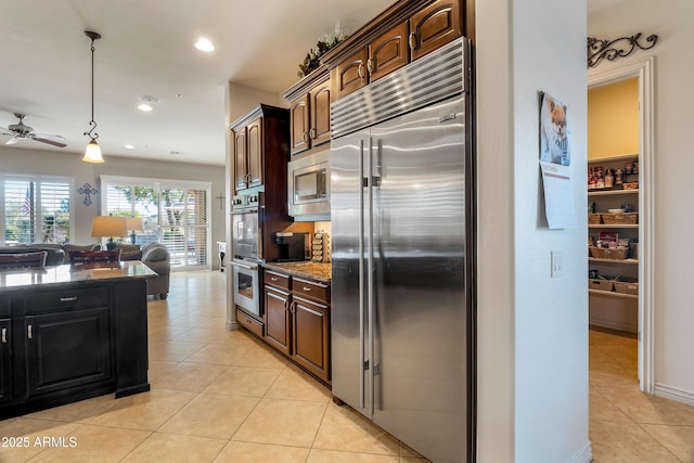 kitchen with light tile patterned floors, ceiling fan, dark stone counters, built in appliances, and pendant lighting