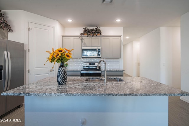 kitchen featuring sink, stainless steel appliances, decorative backsplash, light stone counters, and a kitchen island with sink