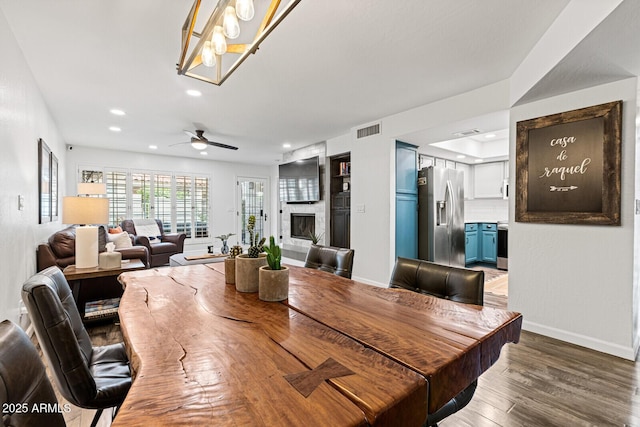 dining area featuring a large fireplace, dark wood-type flooring, and ceiling fan with notable chandelier