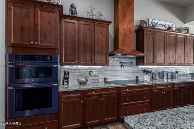 kitchen featuring wall chimney range hood, decorative backsplash, light stone countertops, black electric stovetop, and stainless steel double oven