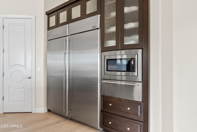 kitchen featuring glass insert cabinets, built in appliances, dark brown cabinetry, a warming drawer, and light wood-style flooring